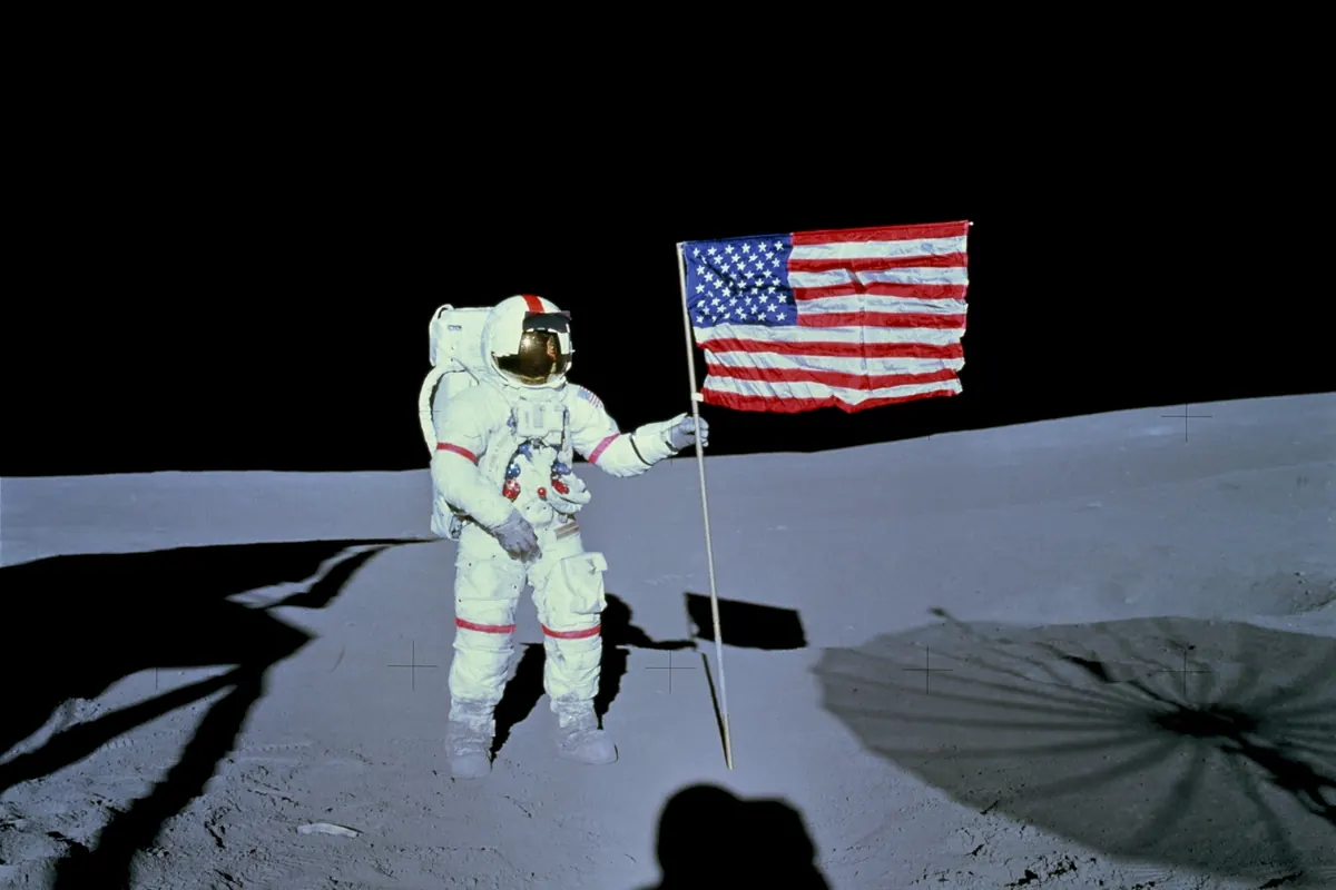 An astronaut in a white spacesuit beside a United States flag on the gray lunar surface