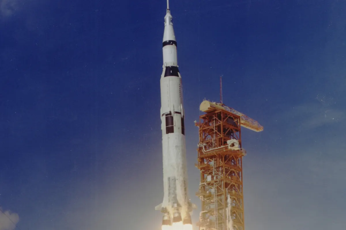 A Saturn V rocket lifting off against a deep blue sky with flame and smoke below it