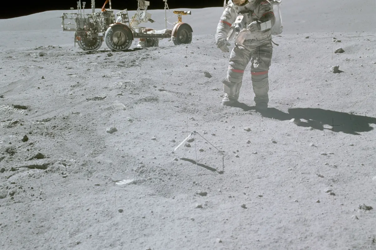 An astronaut in a dusty white spacesuit walking on gray Moon soil beside a lunar rover