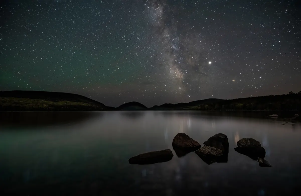 The Milky Way above a still lake and rocks in Acadia National Park
