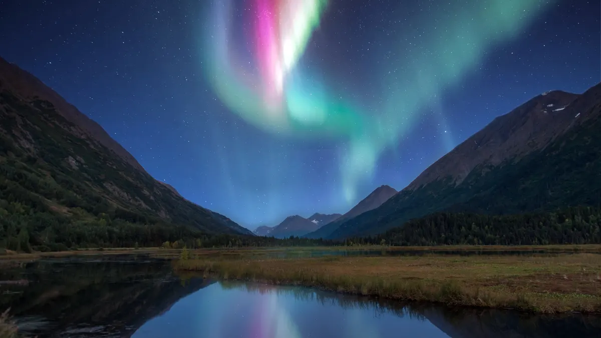 Green and pink northern lights above a mountain valley and lake in Alaska