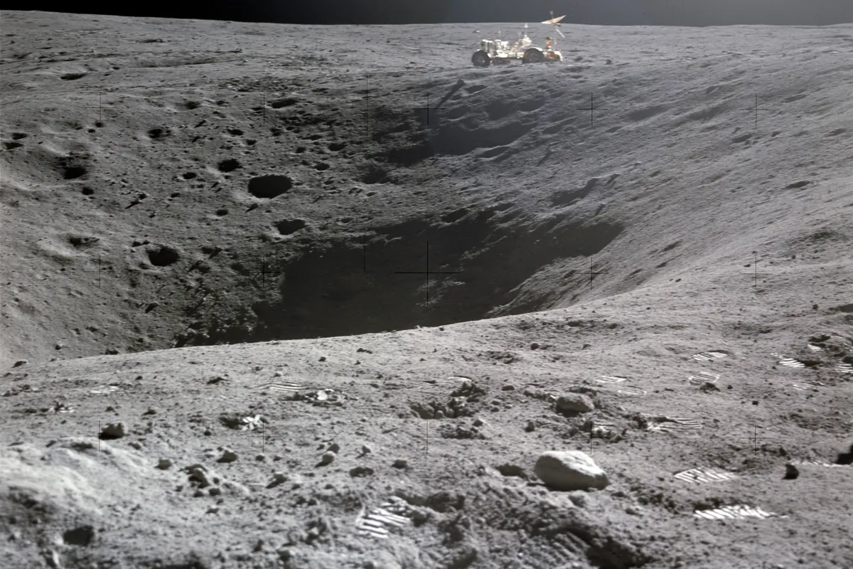 A boulder-strewn crater on the gray lunar surface with the lunar rover parked on the far side