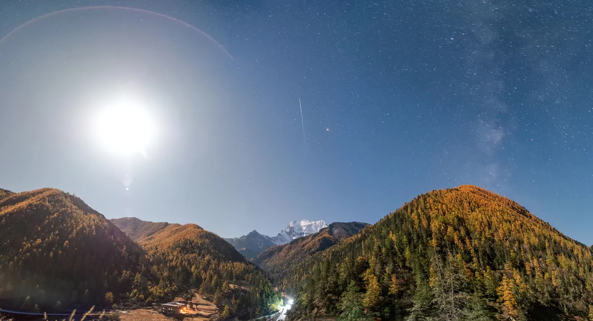 A thin white meteor streak above a moonlit mountain valley with autumn larch forest and a snow-capped peak