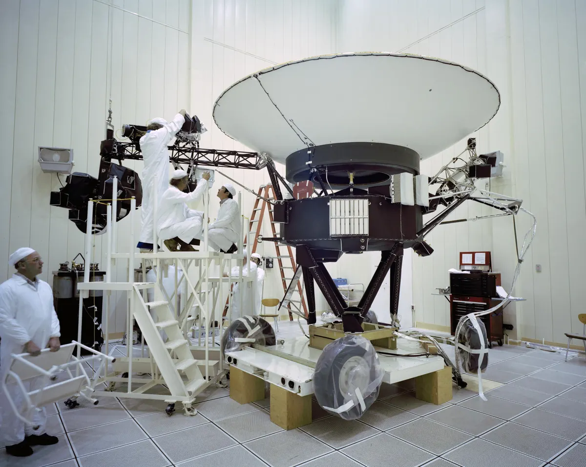 Engineers in white suits working on the Voyager 2 spacecraft and its large dish antenna in a clean room