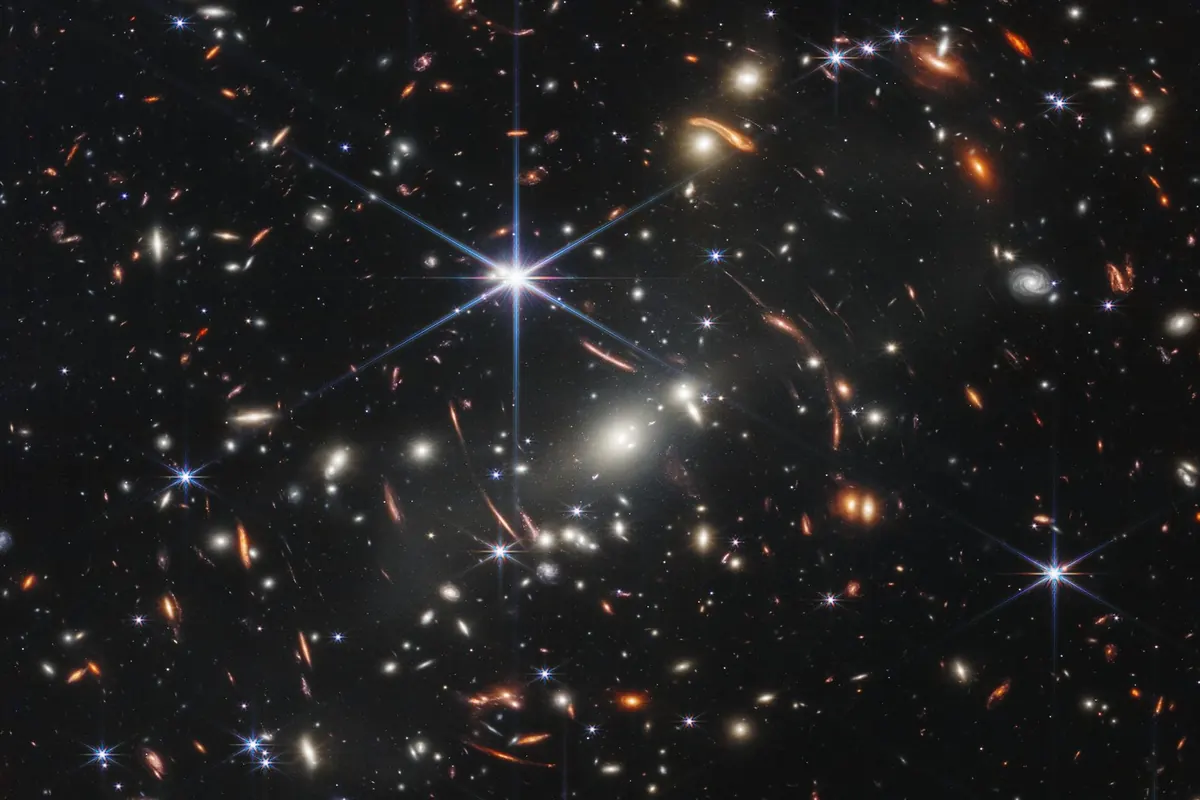 Thousands of galaxies in near-infrared light around a bright star with diffraction spikes, with arcs of lensed galaxies